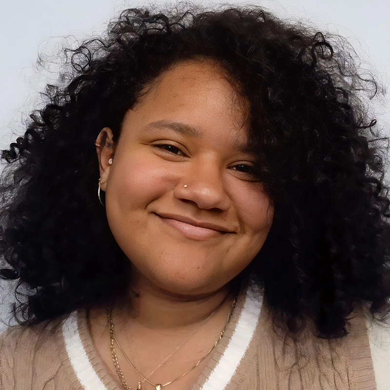 Anjalee Hardy, a young woman with medium brown skin and dark, curly hair, smiles gently at the camera. She wears a beige sweater with a white collar, gold hoop earrings, layered necklaces, and a small nose stud against a light background.