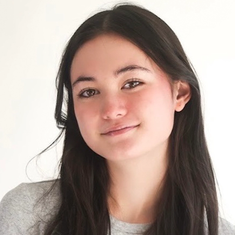 A young woman with long, straight dark hair and fair skin smiles gently. She is wearing a light gray top and is facing the camera with a neutral, light background. Her expression is calm and friendly.