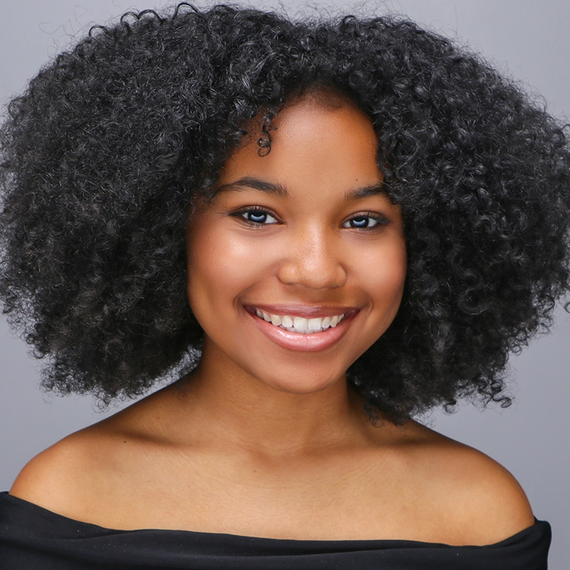 A young woman with medium-dark skin and natural, voluminous, curly black hair smiles brightly. She has clear skin, minimal makeup, and is wearing an off-the-shoulder black top against a plain gray background.