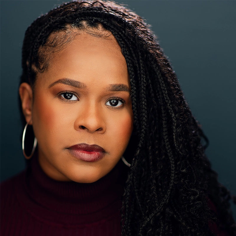 Madison Keenoy, with medium-dark skin and long, braided black hair, wears large hoop earrings and a burgundy turtleneck. She gazes seriously at the camera, her natural makeup highlighting defined brows and dark lipstick against a blurred backdrop.