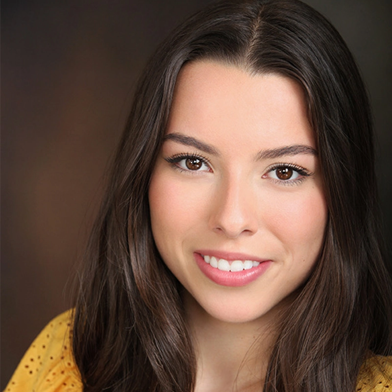 A young woman with long brown hair and brown eyes smiles at the camera. She is wearing a yellow top and has natural makeup. The softly blurred background in brown tones creates a warm atmosphere, reminiscent of Quinton Robinson's style.