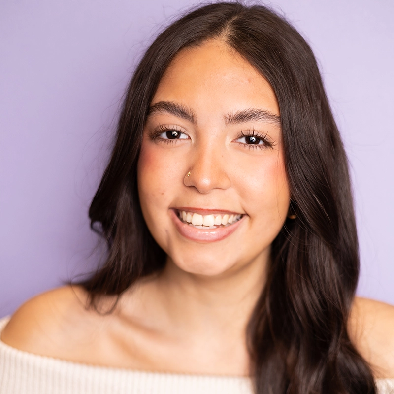 Maya Annunziato, a young woman with long dark brown hair and a nose ring, smiles at the camera. She wears a white off-shoulder top and natural makeup against a smooth, light purple background.