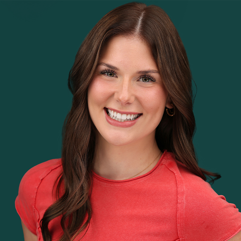 Bailee Loging, a young woman with long brown hair and light skin, smiles widely, showing teeth. She wears a coral short-sleeve top and gold hoop earrings against a solid dark teal background. Her loose waves frame her face as she faces the camera.