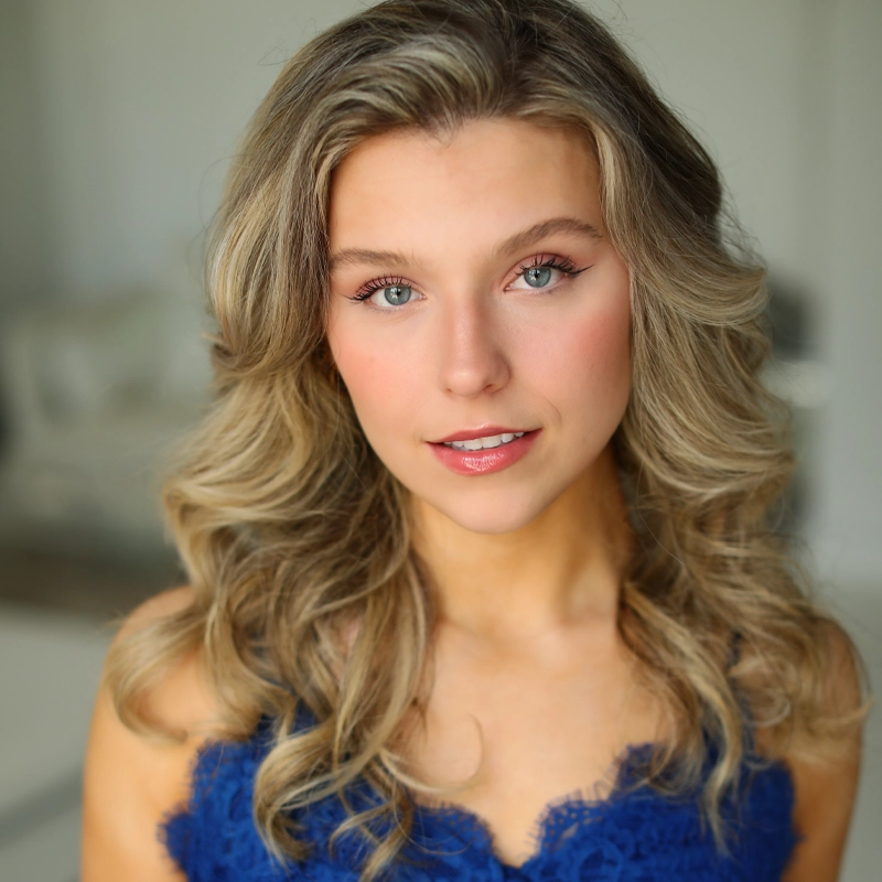 A young woman with wavy, blonde hair and blue eyes smiles softly. She wears a cobalt blue lace top and natural makeup, standing in a softly lit, blurred indoor setting. Her expression is calm and welcoming.