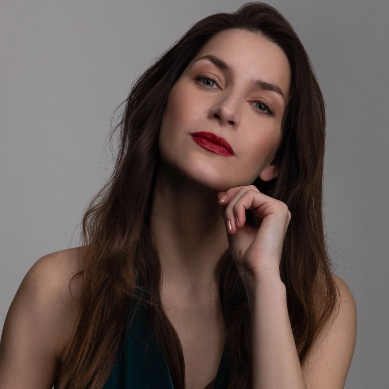 Martina Barta, with long brown hair, poses against a plain gray background in a dark sleeveless top. She looks confidently at the camera, head tilted and chin resting on her hand, wearing bold red lipstick and natural makeup.