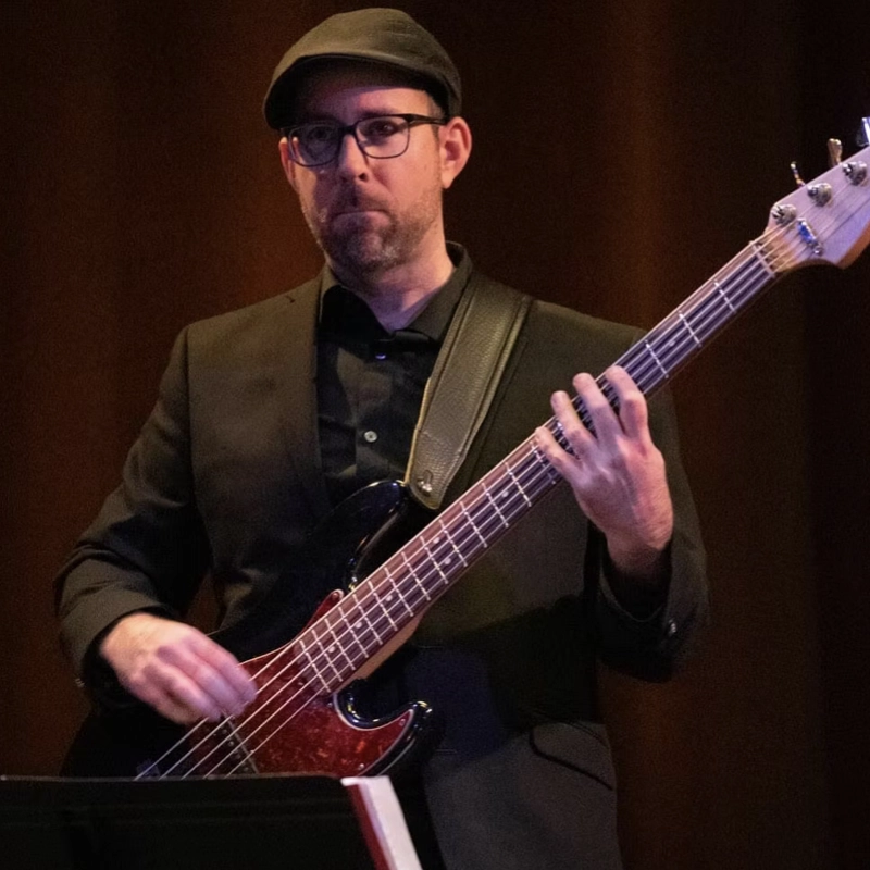 A man wearing glasses, a black flat cap, and a black suit plays an electric bass guitar on stage. He reads from sheet music in front of him, focused on his performance. The background is dark brown and softly lit.