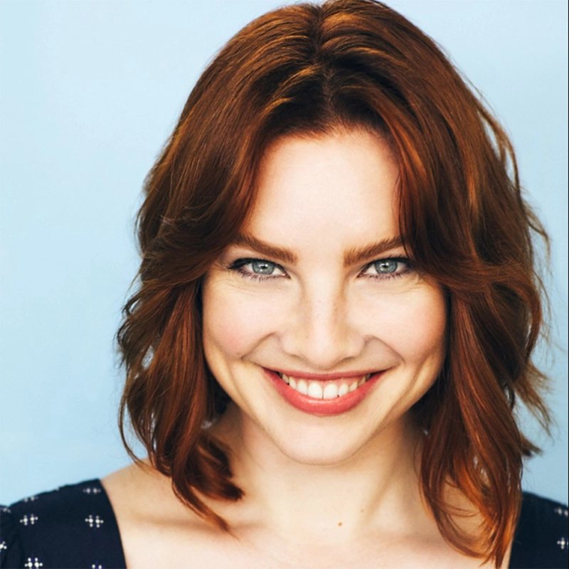 A headshot of Ashley Wells, a smiling woman with shoulder-length auburn hair, blue eyes, and a dark top with a white pattern set against a light blue background.