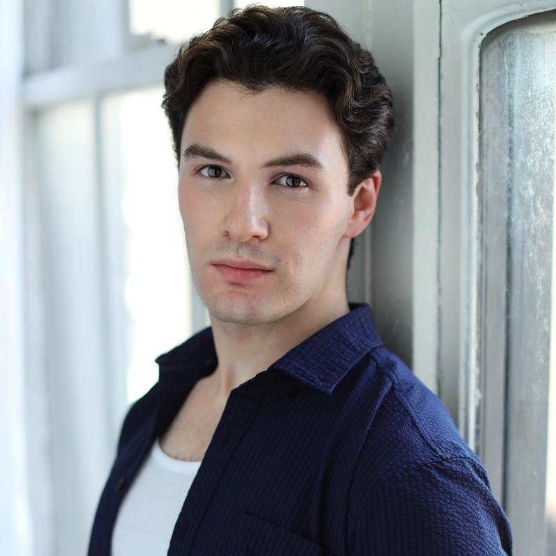 A young man with short, wavy dark hair and fair skin stands indoors by a window. Wearing a navy blue textured shirt over a white top, Elliot Block gazes softly at the camera as natural light illuminates his face. The background is mostly out of focus.