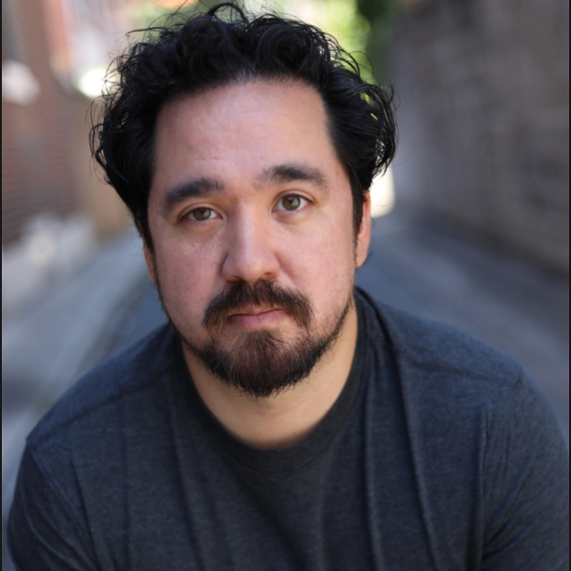 A man with wavy dark hair, a mustache, and beard looks directly at the camera with a neutral expression. He wears a dark gray t-shirt and sits outdoors in a blurred alleyway with brick and stone walls in the background.