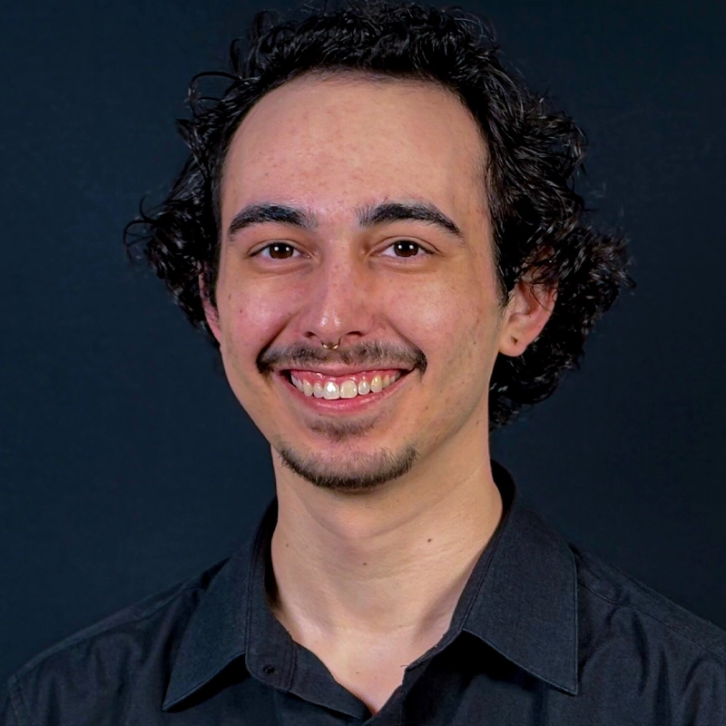 A young adult with curly dark hair, a trimmed mustache and goatee, and light skin smiles at the camera. Trevor P. Bourland is wearing a dark button-up shirt and is posed in front of a dark, solid background.