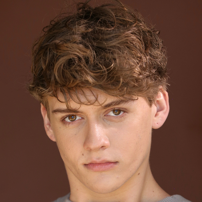 A young person with tousled light brown hair and hazel eyes, possibly Coral Rosario-Laboy, stares directly at the camera. They have fair skin, a neutral expression, and wear a light top against a plain dark background. The face remains the central focus.