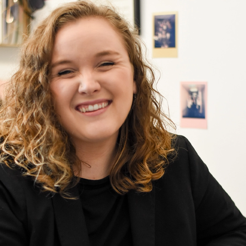 Kerrianne Brennan, a young woman with curly blonde hair, smiles warmly at the camera. She is wearing a black top and black blazer. The background is slightly blurred, with framed photos and a white wall visible behind her.