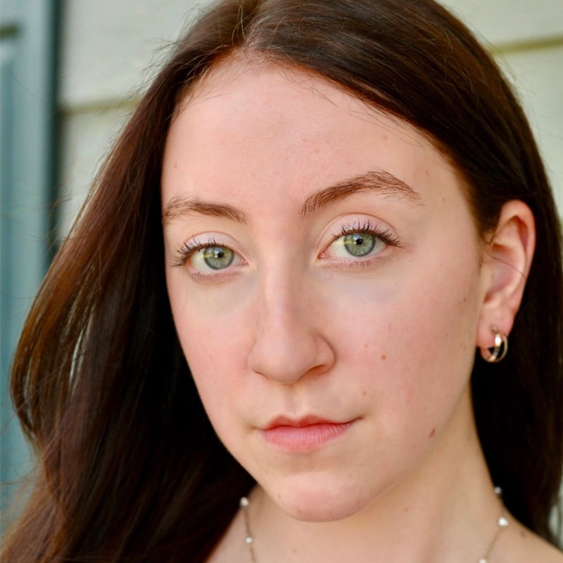 A young woman with long brown hair and green eyes looks directly at the camera. She has light skin, natural makeup, and wears small hoop earrings. The softly blurred background suggests Leigh Renken standing by light-colored siding and a window frame.