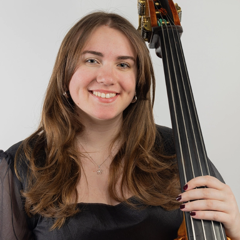 Ashleigh Budlong, a young woman with long brown hair, smiles at the camera while holding the neck of a double bass. She wears a sheer black top, a delicate necklace, and dark nail polish against a plain white background.
