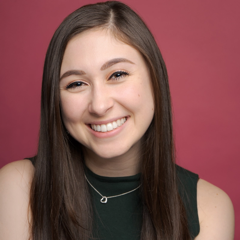 Sydney Burtis, a young woman with long straight brown hair, smiles warmly at the camera. She has fair skin and wears a sleeveless dark green top with a silver heart-shaped necklace, set against a solid reddish-pink background.