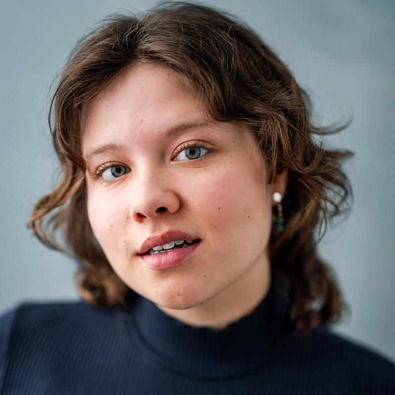 A young person with wavy brown hair and light skin wears a dark ribbed turtleneck and colorful earrings. They have light eyes, natural makeup, and a gentle, neutral expression. The background is soft blue and out of focus.