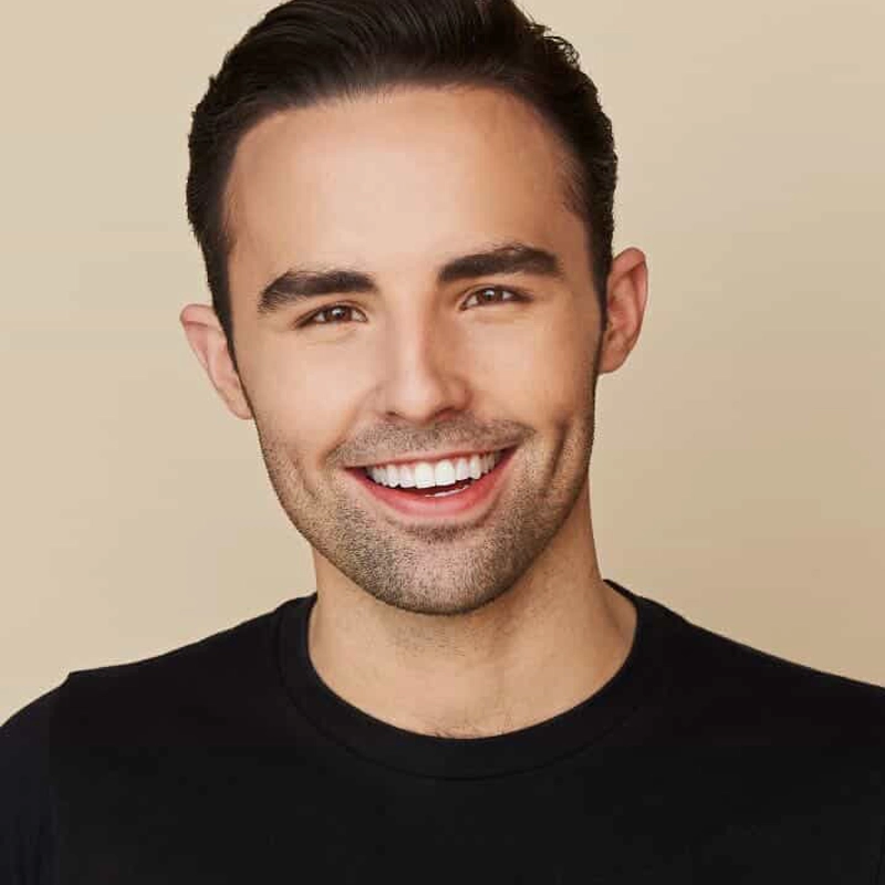 A young man with fair skin and dark hair, styled neatly, smiles broadly at the camera. Jack Cahill-Lemme wears a plain black crew-neck shirt and stands against a light beige background. His teeth are very white, and his complexion is clear.