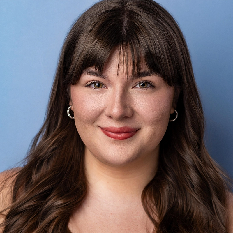 Anjalee Hardy, a young woman with long, wavy brown hair and bangs, smiles softly. She wears silver hoop earrings and natural makeup with pink lipstick. The solid light blue background creates a clean, professional headshot look.