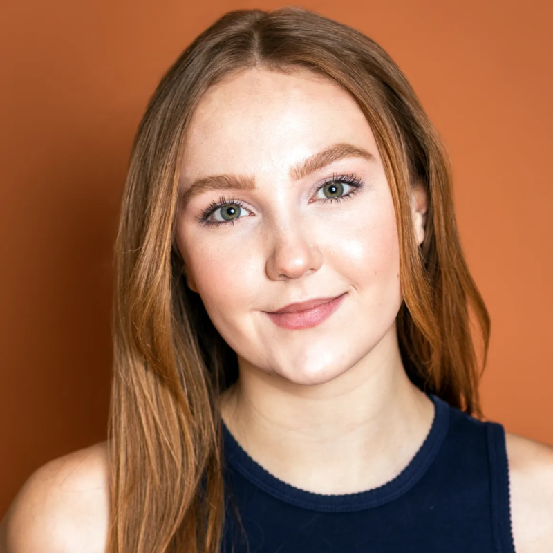 A young woman with fair skin, long straight auburn hair, and green eyes smiles softly at the camera. She wears a sleeveless navy top and is posed in front of an orange-brown background. Her expression is relaxed and confident.