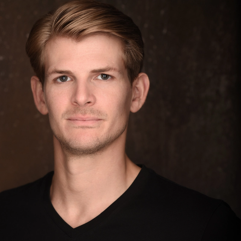 A young man with light skin and short, neatly styled blonde hair looks at the camera with a slight smile. He wears a black V-neck shirt and stands against a dark, softly textured background. The lighting highlights his facial features.