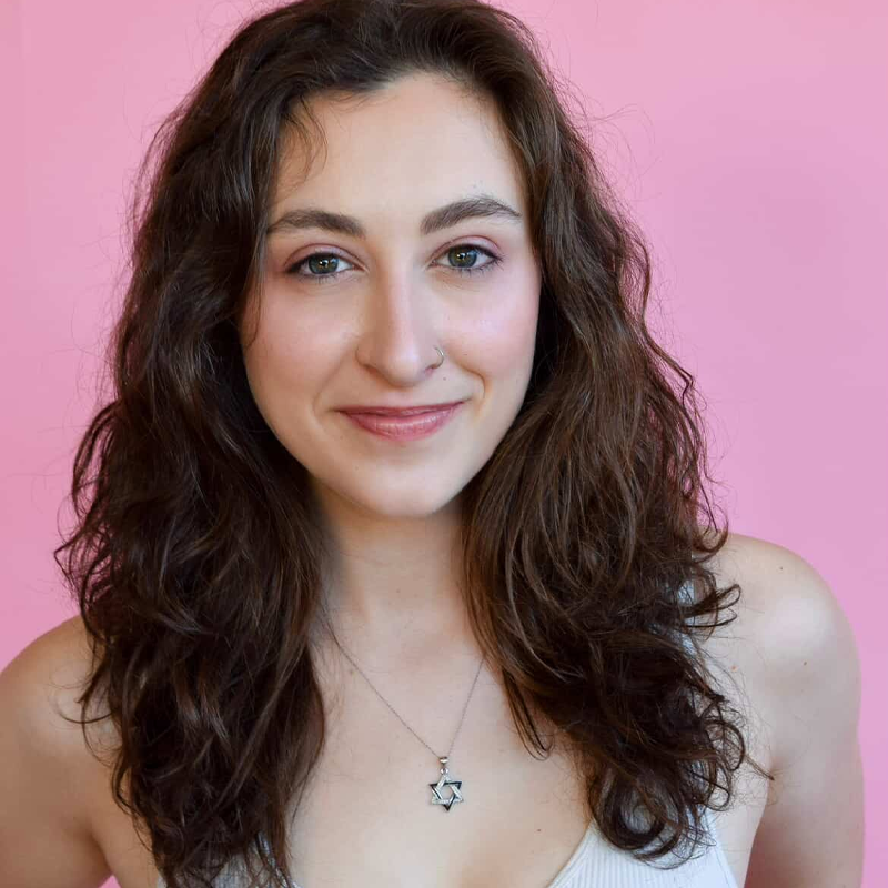 Sophia Carvalho, a young woman with long, wavy brown hair and fair skin, stands in front of a pink background. She smiles softly, wearing a white tank top and a silver necklace with a Star of David pendant.
