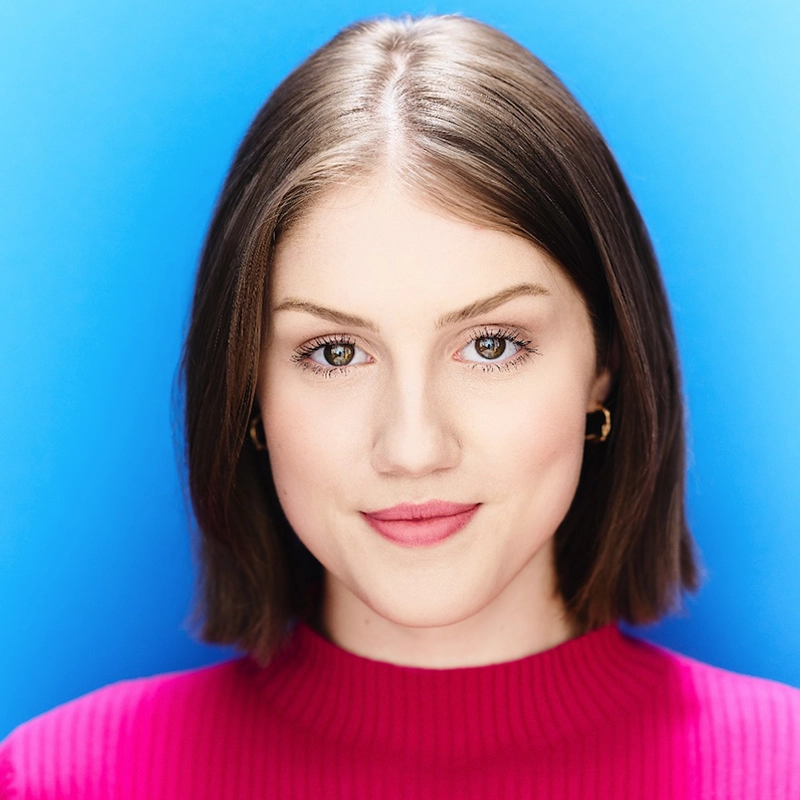 A young woman with fair skin and short brown hair, Madison Keenoy wears a bright pink ribbed top and small gold hoop earrings, smiling softly in front of a solid bright blue background. The portrait is well-lit and she faces the camera directly.
