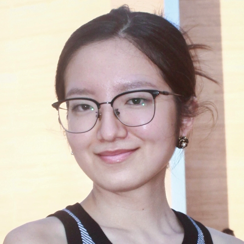 A young woman with fair skin, dark hair tied back, and wearing glasses smiles gently. She has stud earrings and wears a sleeveless black top with white trim. The softly lit background hints at Spencer Lawson's refined, contemporary style.