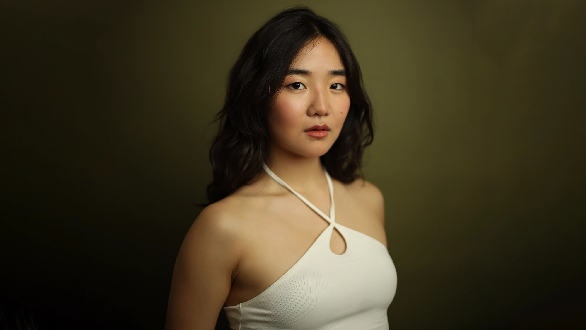 Claire Kwon, sporting a white halter top with a keyhole cutout, stands confidently before an olive-green background. Her wavy dark hair frames her neutral expression and soft, natural makeup in this Claire Squared portrait.