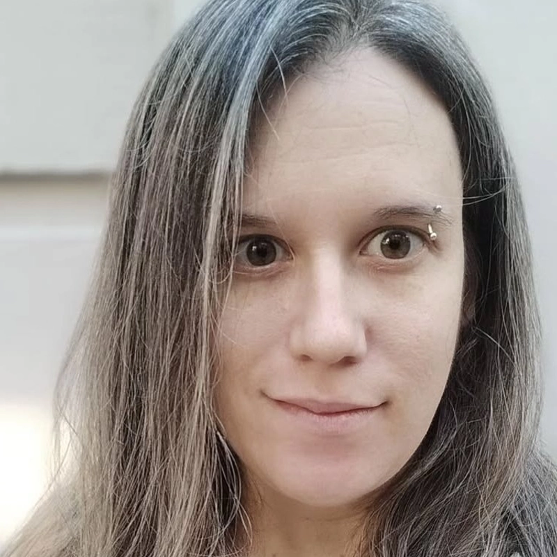 Caitlin Coleman, a woman with fair skin and straight, shoulder-length brown hair with gray streaks, looks at the camera with a neutral expression. She has a silver eyebrow piercing above her right eye and a softly blurred light beige background.