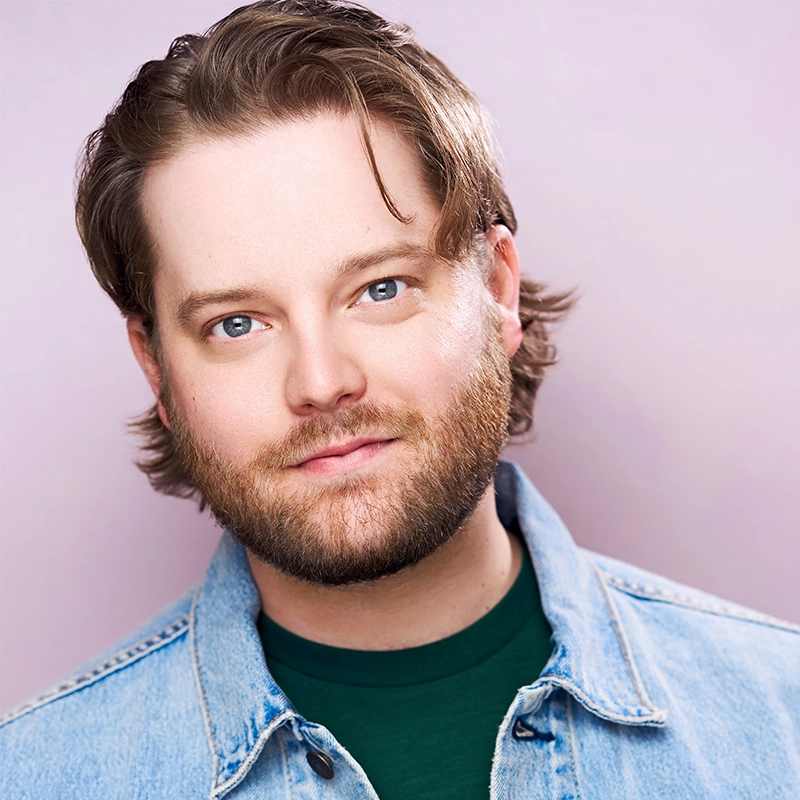 Headshot of Ashley Wells, a man with a beard wearing a green t-shirt under a denim jacket. He is looking directly at the camera with a neutral expression.