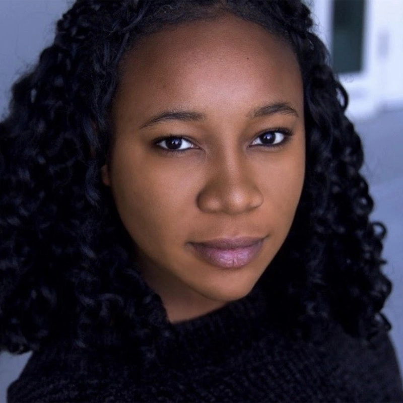 Aisha Collins, a young woman with curly black hair and medium-brown skin, looks directly at the camera with a gentle, confident expression. She wears a black textured top against a blurred, cool-toned indoor or urban background.