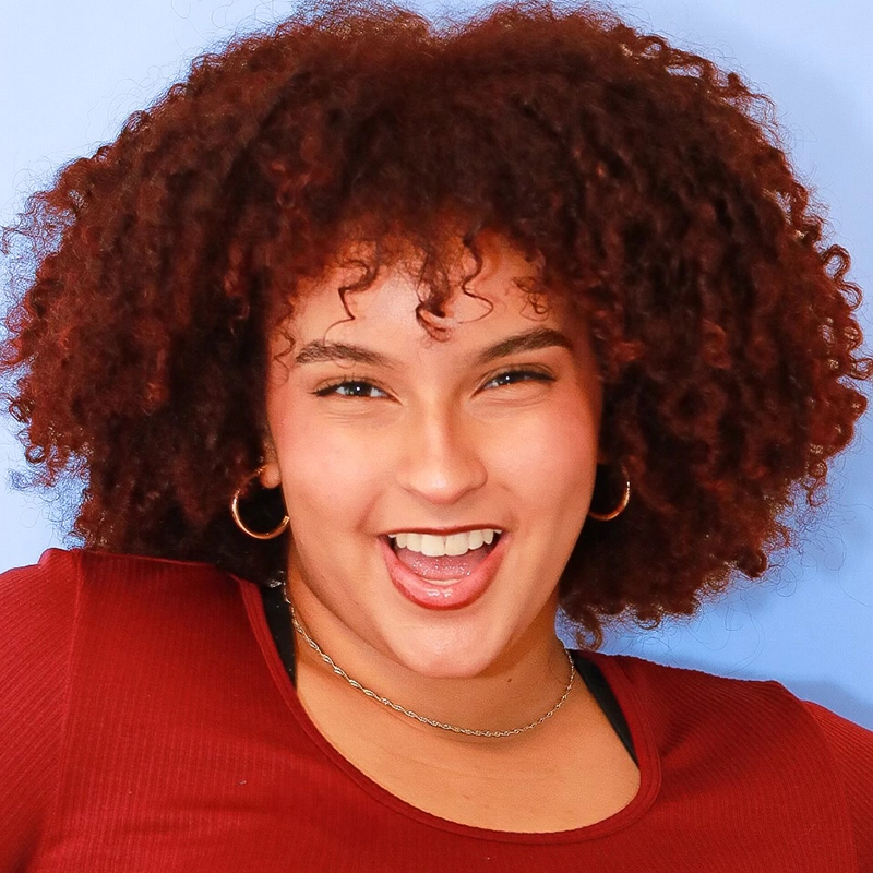 Coral Rosario-Laboy, a young woman with curly, voluminous reddish-brown hair, smiles widely with her mouth open. She wears gold hoop earrings, layered necklaces, and a maroon ribbed top against a solid light blue background.