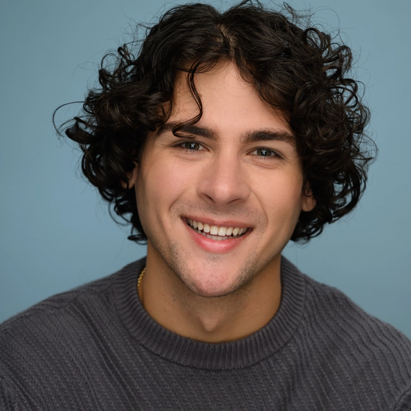 A young adult with curly dark hair and light skin, Donovan Counts, smiles warmly at the camera. He is wearing a textured, dark gray sweater and sits in front of a plain, soft blue background. The lighting highlights his friendly expression.