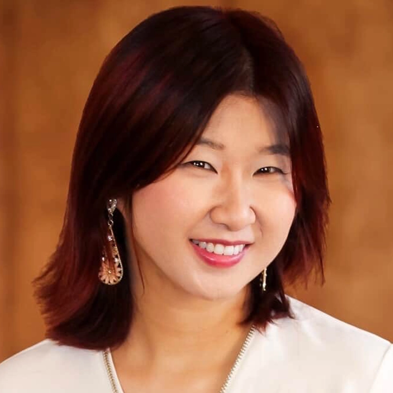 A woman with straight, shoulder-length dark hair and wearing a white top smiles at the camera. She has drop earrings, and her relaxed pose is set against a softly blurred, warm brown background by Spencer Lawson.