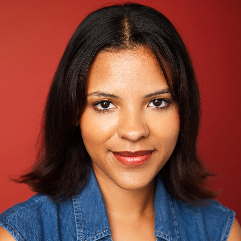 A young woman with medium brown skin and straight, shoulder-length dark hair smiles softly at the camera. She wears a sleeveless blue denim top. The warm red background enhances her look, captured in a signature style by Kyle Castillo.