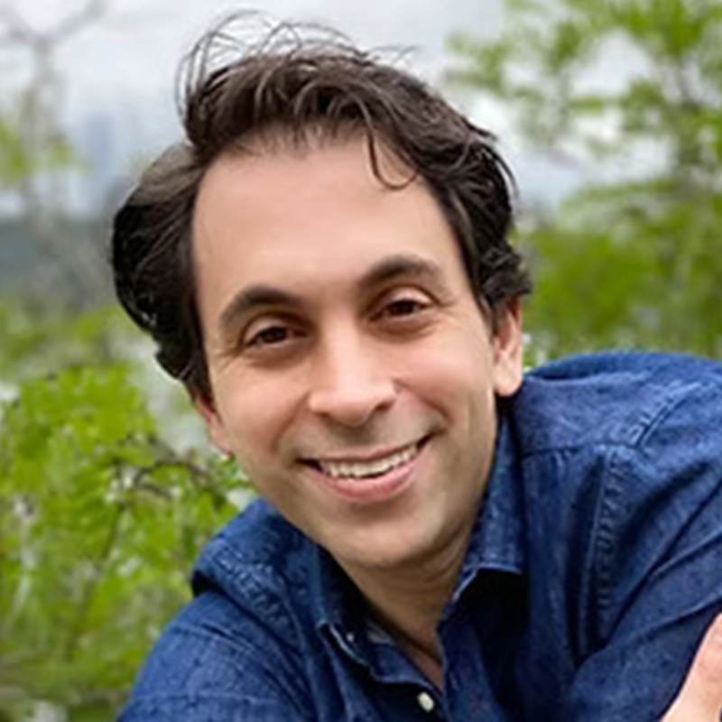 Ashley Wells, a smiling man with dark hair, wears a blue collared shirt outdoors with soft-focus greenery in the background.