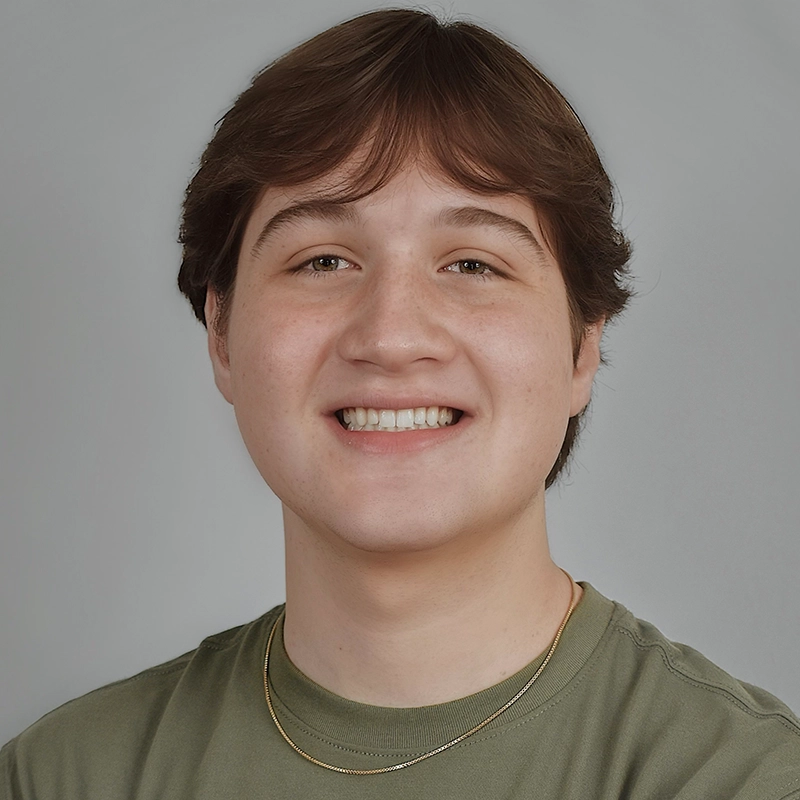 A young person with short brown hair smiles at the camera. Gabe Woodard is wearing a light green T-shirt and a thin gold chain necklace. The plain, light gray background keeps the focus on their friendly expression.