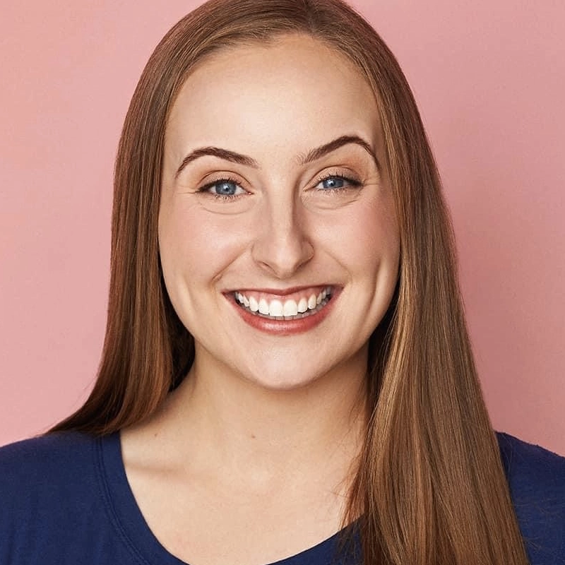 Laura Dellis, a young woman with fair skin, straight light brown hair, and blue eyes, smiles widely to reveal her teeth. She wears a blue top and is posed against a solid light pink background under even, warm lighting.