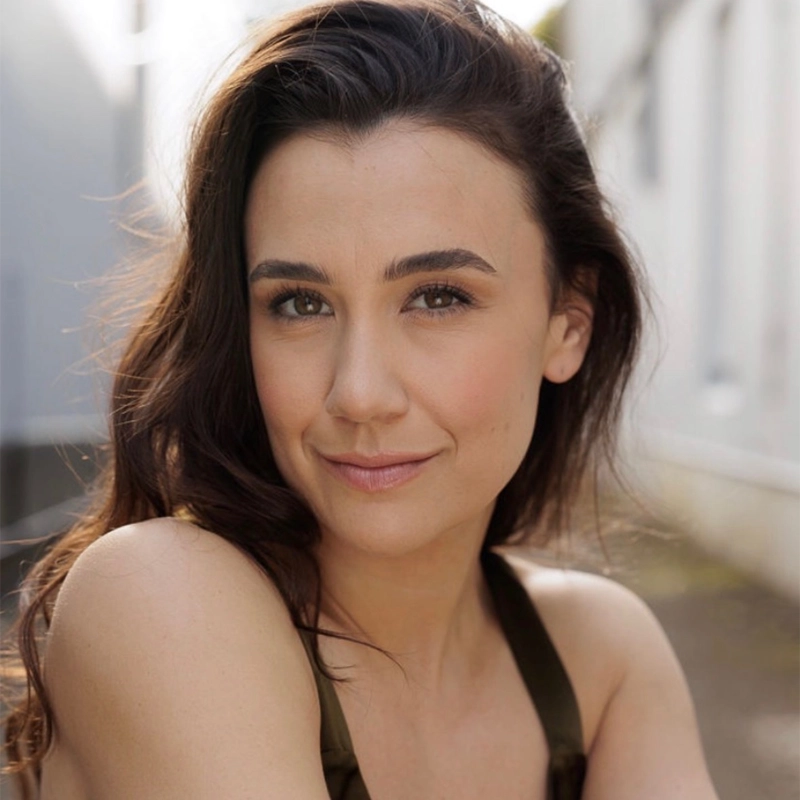Samantha Dodemaide, with long brown hair and light skin, smiles softly at the camera. She wears a sleeveless top and sits outdoors in natural sunlight, with a blurred backdrop of white buildings and a calm, relaxed expression.