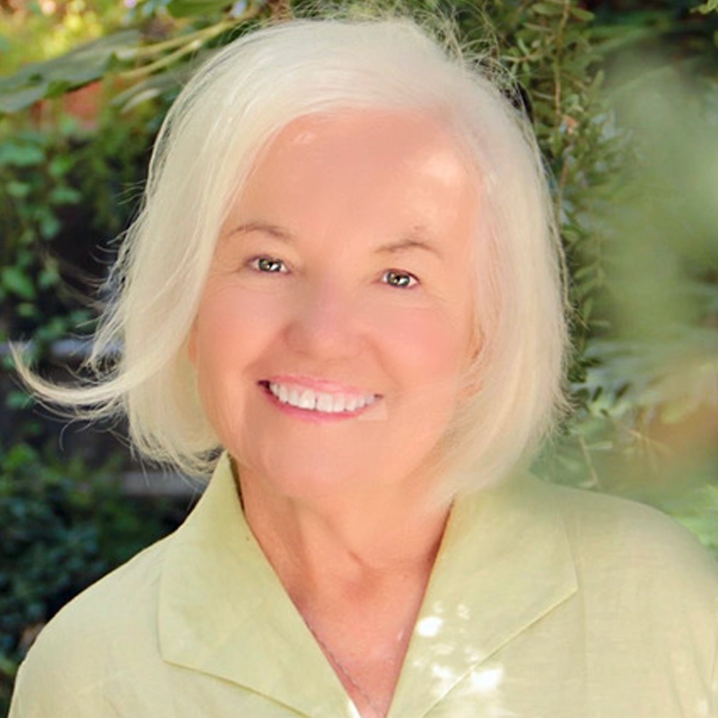 An elderly woman with short, white hair smiles warmly at the camera. Jamie Donnelly wears a light green collared shirt, surrounded by greenery in an outdoor setting. Sunlight softly illuminates her face, creating a peaceful and cheerful mood.