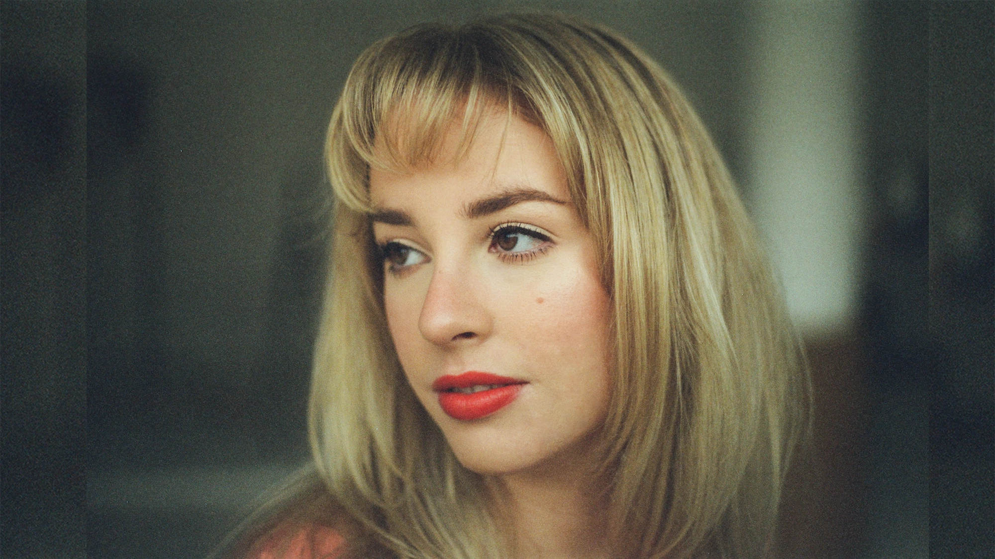 A woman with long blonde hair and bangs looks slightly to her left. She has fair skin, red lipstick, subtle eye makeup, and a small beauty mark on her cheek—reminiscent of Ella Mae Dixon's elegance in classic swingin’ showtunes. The background is softly blurred.