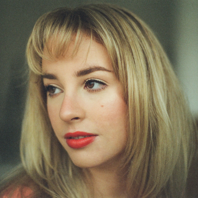 A young woman with light skin, straight blonde hair, and bangs gazes thoughtfully to the left. She has red lipstick, subtle eye makeup, and a small beauty mark on her cheek. The background is soft and out of focus, highlighting her face.