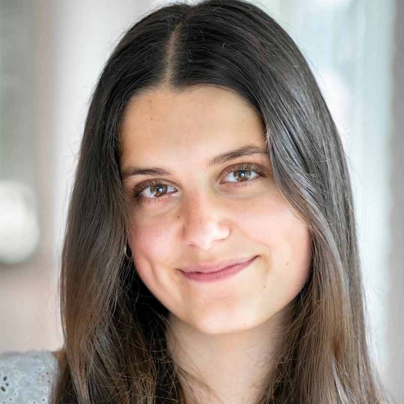 A young woman with long, straight brown hair and blue-green eyes smiles gently at the camera. She has light skin, subtle makeup with a hint of coral on her lips, and is wearing a white top. The softly blurred background suggests natural indoor light.