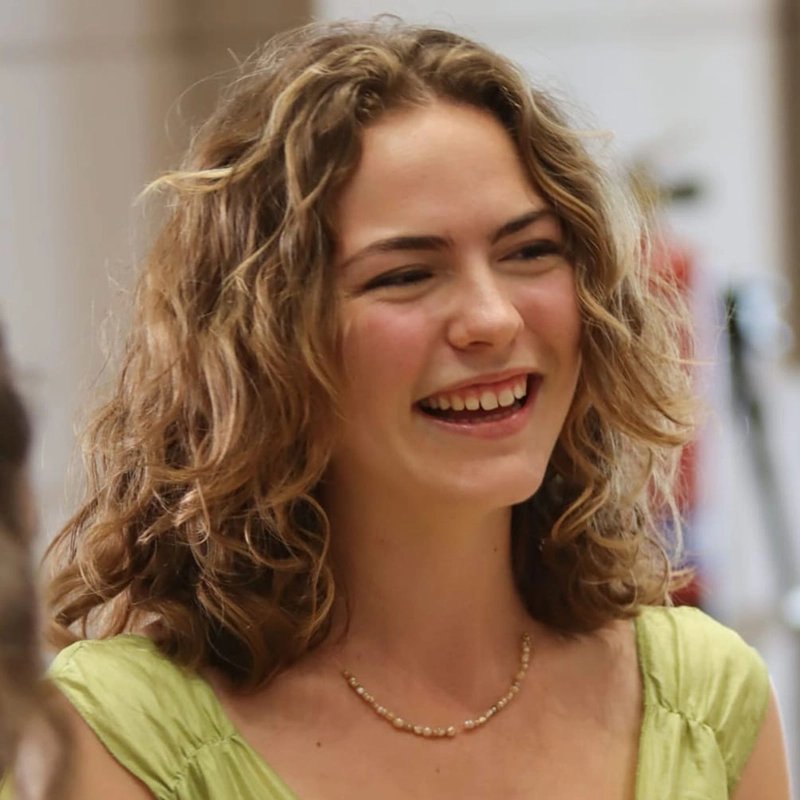 A young woman with curly light brown hair smiles brightly. Elsa Rose McIntyre wears a light green top and a delicate beaded necklace. The softly blurred background hints at an indoor setting in Córdoba. Her expression is joyful and relaxed.