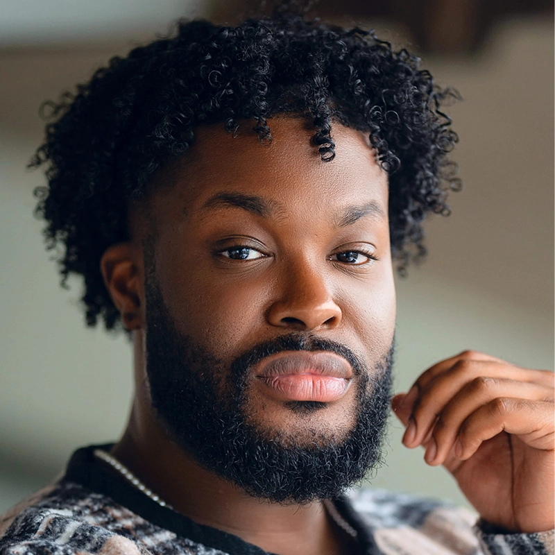 A headshot of Ashley Wells, a Black man with a full beard and short, curly hair, looking directly at the camera with one hand raised toward his chin.