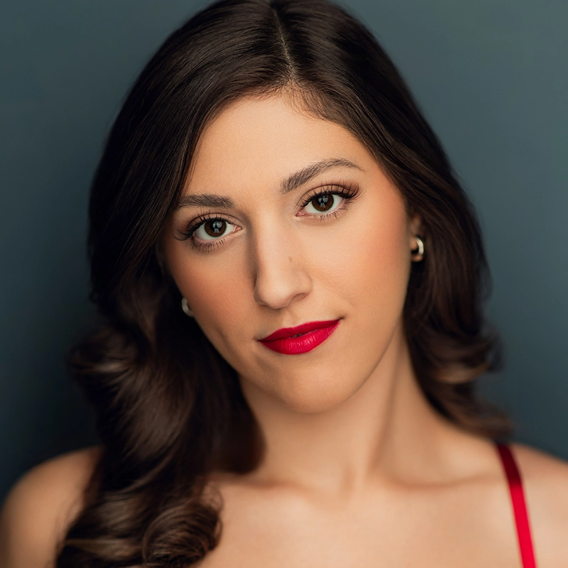 Grace Whetstone, a young woman with long, wavy brown hair, poses against a dark blue background. She wears bold red lipstick, pearl earrings, and a red spaghetti strap top, gazing directly at the camera with a neutral expression.
