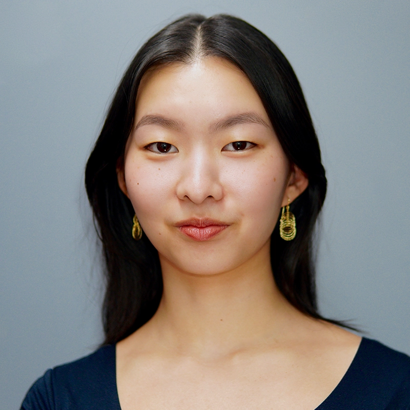 A young woman with long, straight black hair and light skin smiles gently at the camera. She wears gold drop earrings and a dark navy blue top. The background is plain, light gray, creating a simple and clean portrait setup.