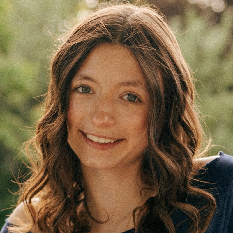 A young woman with wavy brown hair and green eyes smiles at the camera. She is outdoors, surrounded by greenery and sunlight. Wearing a dark top, her hair falls loosely over her shoulders in this Quinton Robinson-inspired portrait.