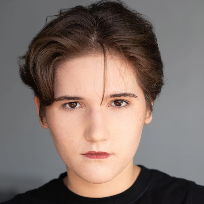 Madison Keenoy, with short brown hair, fair skin, and brown eyes, stares directly at the camera with a neutral expression. She wears a black top against a plain gray background, one lock of hair falling over her forehead.