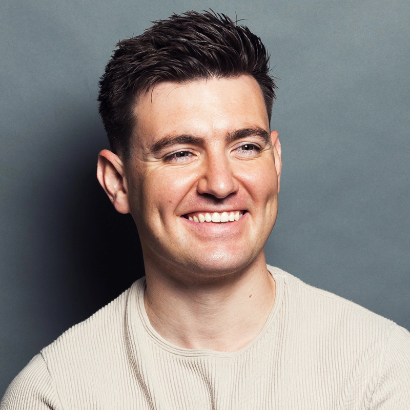 A smiling man with short dark hair, wearing a light beige ribbed sweater, poses against a plain gray background. He is facing slightly to the right, showing his teeth as he smiles warmly. The lighting is even, highlighting his cheerful expression.